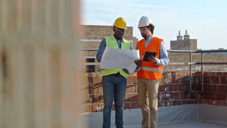 African-American-and-Caucasian-men-constructors-in-hardhats-walking-slowly-at-the-building-site-with-drafts-of-plan-and-rablet-device-while-talking-and-discussing-next-parts-of-work.